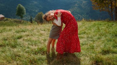 Happy woman with daughter hugging on green meadow sunny day. Smiling mother with lovely child enjoying summer holiday in mountains. Cheerful mom with cute girl spending time together outdoors.