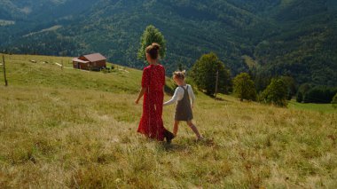 Back view of young woman walking with daughter on green hill. Unknown mother looking at mountains panorama with little girl outdoors. Lovely child with mom enjoying summer vacation in mountains.