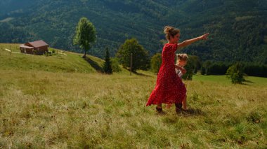 Happy mother walking with daughter on green grass mountain hill. Carefree woman pointing finger at nature views outdoors. Beautiful mom spending summer holiday with lovely child. Leisure together.