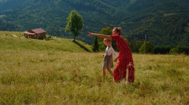 Relaxed young woman with girl enjoy walking on meadow summer holiday. Happy mother with daughter resting together in mountains. Carefree mom pointing finger at beautiful nature views outdoors.