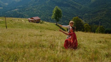 Carefree mother sitting near daughter on meadow sunny day. Happy woman relaxing with cute girl in mountains enjoying summer vacation. Young mom in red dress pointing finger at beautiful nature views.