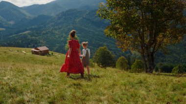 Back view of lovely mother with daughter going down green hill. Unknown woman with cute girl holding hands walking green grass in mountains. Young mom spending family holiday outdoors with child.