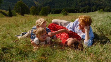 Relaxed family lying on green grass beautiful slope sunny day. Positive young parents resting with children on summer vacation. Smiling couple with kids feeling happy together on mountain hill.