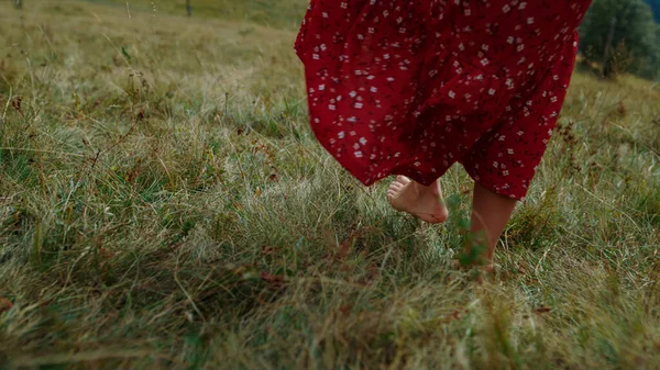 Unknown barefoot girl walking on green grass cloudy day close up. Bare woman feet making steps on meadow summer day. Unrecognizable young lady in red dress enjoying vacation in beautiful mountains.