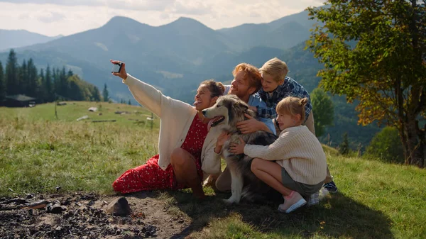 Joyful family taking selfie with husky on cellphone sitting mountain hill summer day. Smiling woman holding smartphone in hand making happy photo. Cheerful twochildren couple with dog posing on nature