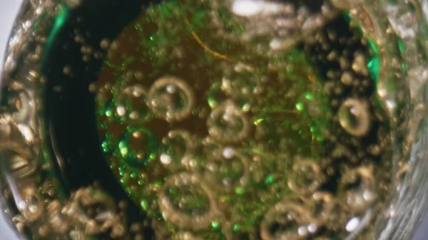 Lager falling splashing flask closeup. Fizzy cold beverage in transparent green bottle. Unfiltered ale splattering different directions in slow motion. Golden alcohol drink overflowing top view