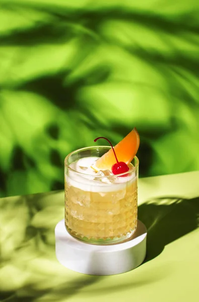 Whiskey sour trendy alcoholic cocktail with bourbon, lemon juice, egg white and ice, rocks glass on bright green background with hard light. Copy space