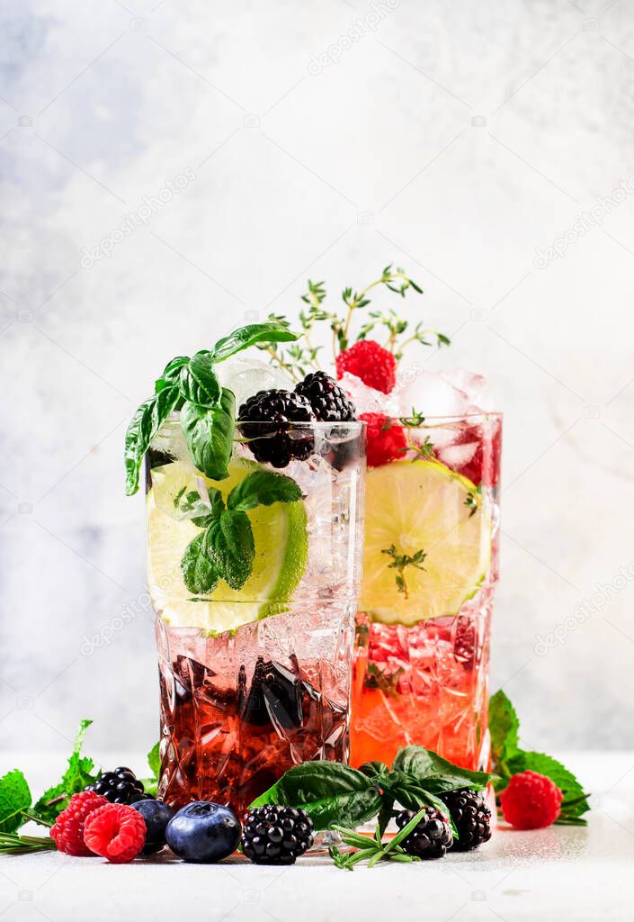 Cocktails or mocktails cold drinks. Classic summer refreshing long drink in highballs with berries, lime, herbs and ice on gray bar counter background