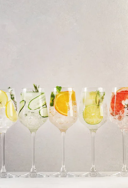 Gin and tonic cocktails set. Trendy Alcoholic drinks with lime, lemon, grapefruit, orange, cucumber, soda and spicy herbs in wine glasses, gray background. Summer cocktail party