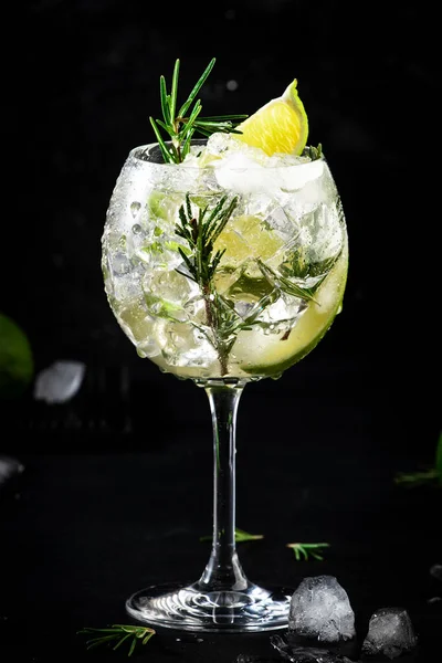 Gin tonic alcoholic cocktail drink with dry gin, rosemary, tonic, lime and ice cubes in wine glass. Black bar counter background, bar tools, copy space