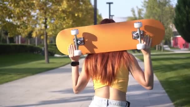Junge attraktive rothaarige Frau mit Skateboard im Park im Freien. Frau mit orangenem Skateboard an einem warmen Sommertag — Stockvideo