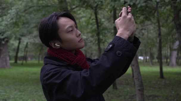 Junger Asiate macht Foto oder Video mit Smartphone im Herbstpark. Der Mann fotografiert die Natur am Telefon — Stockvideo
