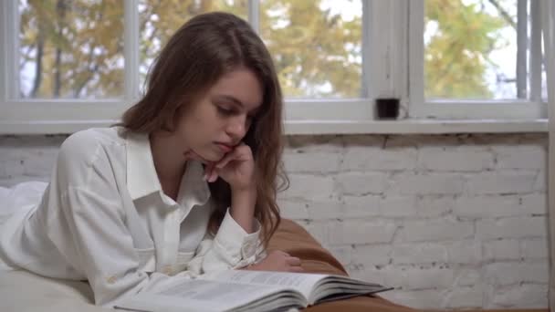 Junge attraktive Frau liest ein Buch auf dem Bett. Schöne Studentin mit einem Buch im Schlafzimmer — Stockvideo