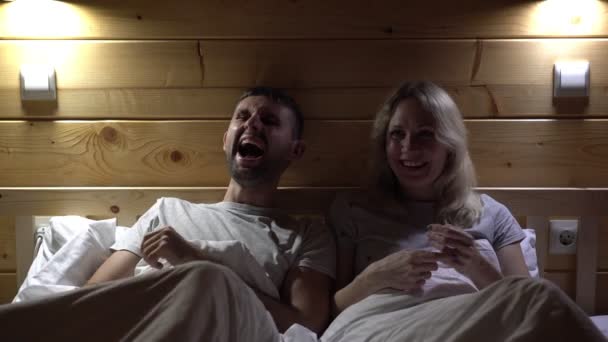 Et ektepar ser på en komedie på sengen. En ung familie ser på tv og ler. – stockvideo