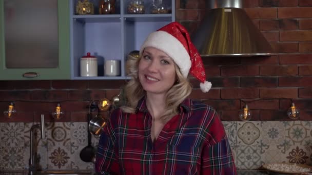 Attraktiv kvinna i köket i tomteluva, julstämning. Porträtt av en lycklig kvinna kvällen före det nya året — Stockvideo