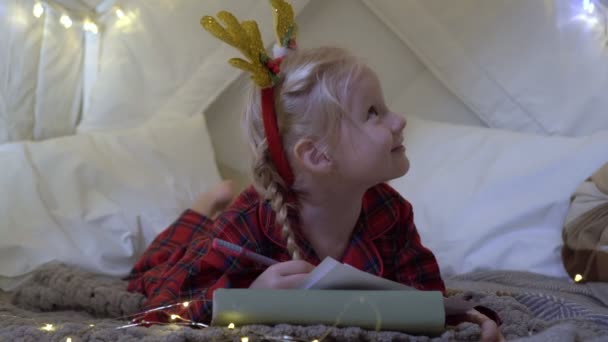クリスマス、子供の女の子は贈り物についてサンタクロースに手紙を書きます。子供は新年への贈り物を夢見ています — ストック動画