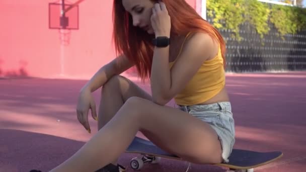 Jovem mulher atraente ouve música com fones de ouvido sem fio. Mulher sexy com skate ao ar livre. Juventude moderna — Vídeo de Stock