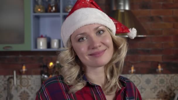 Porträtt av en ung lycklig kvinna i jultomtehatt i julklapp. Kvinnan i köket firar nyår — Stockvideo
