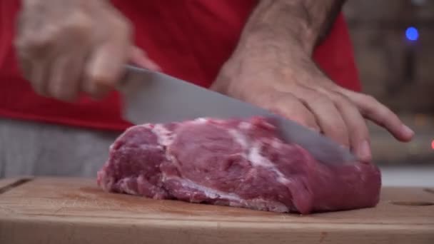 En man skär en bit rått kött med en stor kniv i köket. En man lagar kött hemma — Stockvideo