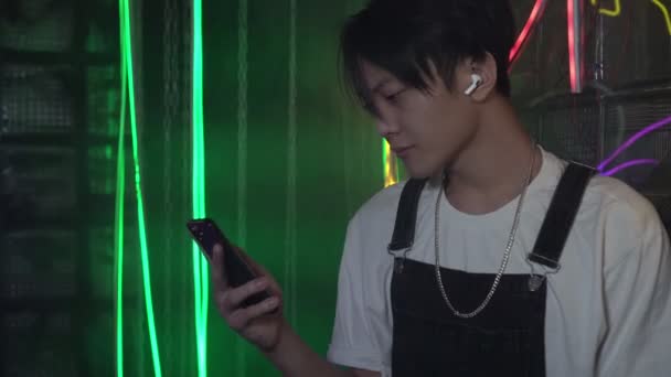 現代のアジア人男性がスマートフォンの画面を見ている。スマートフォンやワイヤレスヘッドフォンを使って音楽を聴く男 — ストック動画