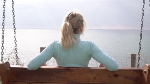 Een vrouw zit op een schommel en kijkt naar de zee. jong gelukkig vrouw kijkt naar de rustig oceaan — Stockvideo