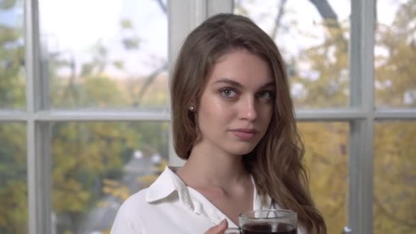 Young woman with a cup of coffee in the morning on the background of the window. Autumn morning, portrait of a young woman in the kitchen. — Stock Video