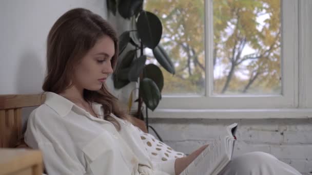 Молода жінка читає книжку на ліжку. Осінній ранок, красива жінка читає книгу вдома — стокове відео