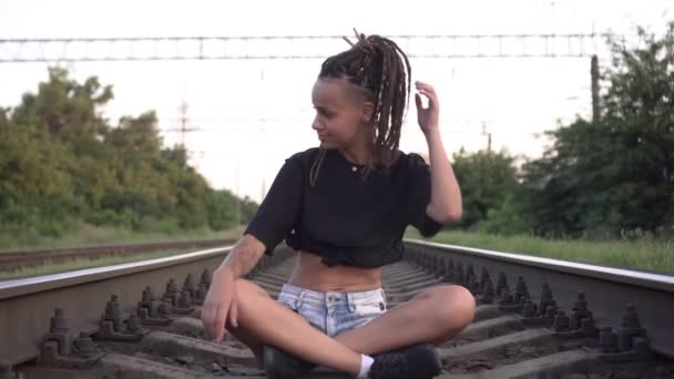 Jovem mulher sexy está sentada nos trilhos do caminho de ferro. Hippie mulher com dreadlocks cabelo — Vídeo de Stock