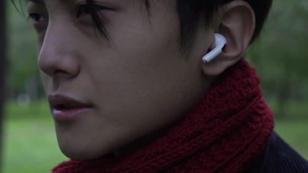Een man luistert naar muziek in een draadloze hoofdtelefoon. Jonge Aziatische man met een koptelefoon buiten. Draadloze oortelefoon in het oor close-up — Stockvideo
