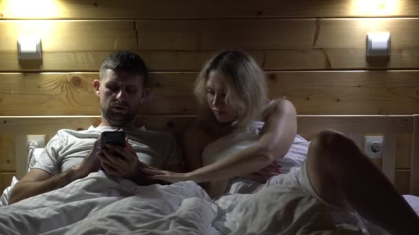 Een man kijkt naar zijn smartphone en wil geen seks met zijn vrouw. Slaperige man wil niet vrijen. — Stockvideo