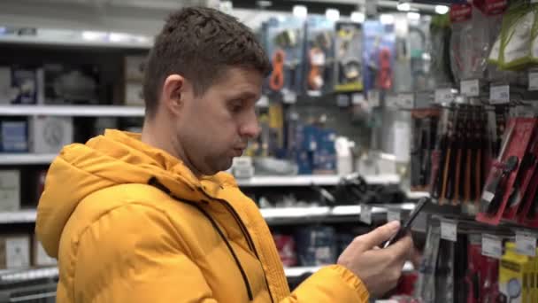 Um homem com um smartphone num supermercado de edifícios. Um homem escolhe um produto numa loja de ferragens — Vídeo de Stock