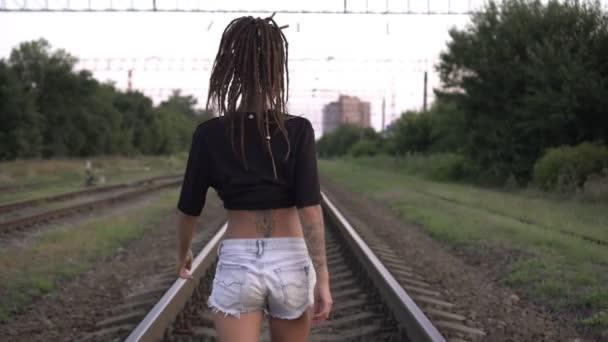 ドレッドロックを持つ若い魅力的な女性が鉄道に沿って歩いています。セクシーな女がレールを歩く。自由と自己表現の概念 — ストック動画