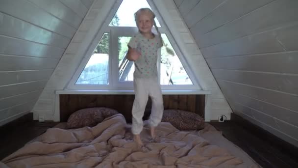 Щаслива дитина стрибає на ліжку в спальні. Весела маленька дівчинка в дерев'яному заміському будинку на горищі — стокове відео