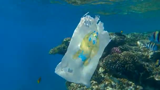 Plastový nákupní taška se žlutým smajlíkem plující pod vodou v blízkosti krásného korálového útesu s tropickými rybami plavat kolem něj. Problém znečištění životního prostředí plastovým odpadem. (4K - 60fps)