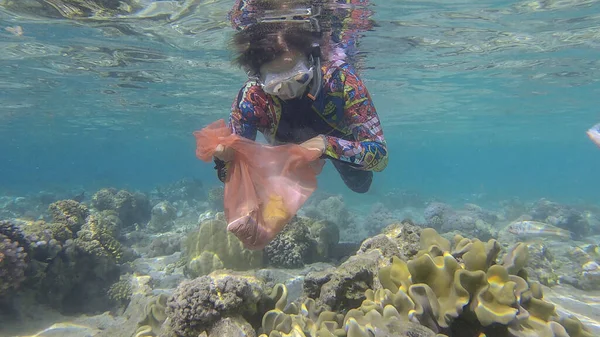 Woman in diving equipment swims and collects plastic debris underwater on the bottom of coral reef. Snorkeler cleaning Ocean from plastic pollution. Plastic pollution of the Ocean. Red sea, Egypt