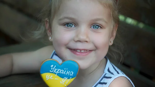 Little blonde girl holds gingerbread in Ukrainian national colors in her hand, it says \