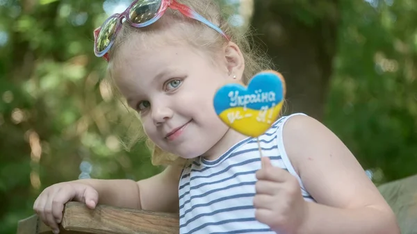 Little blonde girl holds gingerbread in Ukrainian national colors in her hand, it says \