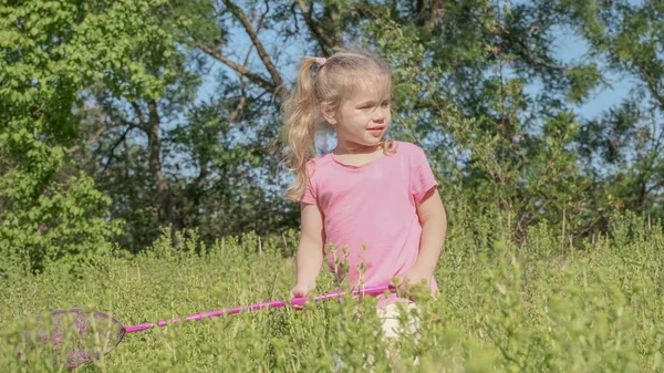 Little girl plays with butterfly net of tall grass in city park. Cute little girl is playing with aerial insect net in meadow on sun day.