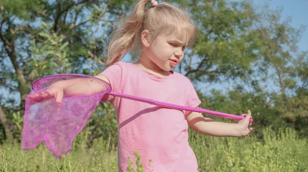 Little girl plays with butterfly net of tall grass in city park. Cute little girl is playing with aerial insect net in meadow on sun day.