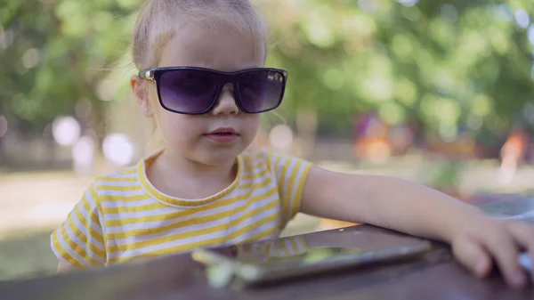 Little girl in mom\'s sunglasses is learning foreign language by repeating words from mobile phone. Close-up portrait of child girl sitting in city park and learning foreign language using cellphone