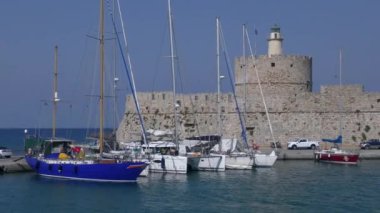 Limanın girişini koruyan antik kule ve rıhtımda duran yatlar, hareket halindeki bir geminin manzarası. Rodos, Yunanistan.