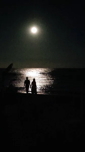 Gecenin bir yarısı deniz manzarası ve dolunay karşısında genç bir adam ve kadının siluetleri. 