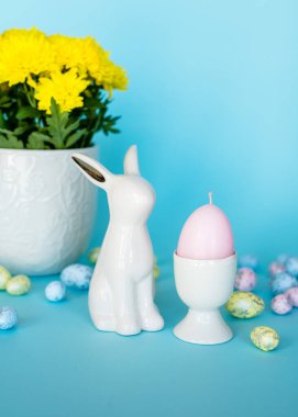 Paskalya kartı. Paskalya porseleni tavşanı, yumurta standında pembe mum yumurtası, mavi arka planda yumurtalar. Pastel renkler. Uzun pankart biçimi