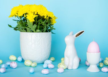 Paskalya kartı. Paskalya porselen tavşanı, boyalı yumurtalar ve mavi arka planda beyaz bir saksıda sarı bir çiçek. Sarı-mavi konsept.