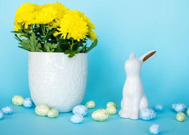 Paskalya kartı. Paskalya porselen tavşanı, boyalı yumurtalar ve mavi arka planda beyaz bir saksıda sarı bir çiçek. Sarı-mavi konsept.