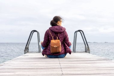 Sırt çantalı genç bir kadın denize bakıyor, ahşap bir iskelede oturuyor. Özgürlük için. Mahremiyet. Adaylık