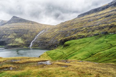 Şelale ve küçük bir köy güzel dağların ve dramatik bulutların arka planına kurulur. Faroe adaları. Danimarka. Avrupa. Manzaralar. Doğa.