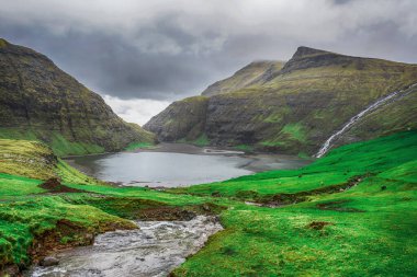 Şelale, güzel dağların arka planına ve dramatik bulutlara karşı deniz koyuna akar. Faroe adaları. Danimarka. Avrupa. Manzaralar. Doğa.
