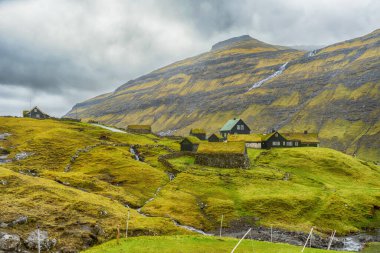 Dağın eteğinde, çim çatılı evleri olan küçük bir köy. Faroe Adaları. Danimarka. Avrupa. Manzaralar. Doğa.