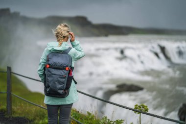 Sırt çantası ve yeşil ceketli sarışın kadın İzlanda 'daki Gullfoss şelalesini ziyaret etti. İzlanda 'nın Altın Daire rotasında ziyaret etmek için ünlü bir yer.